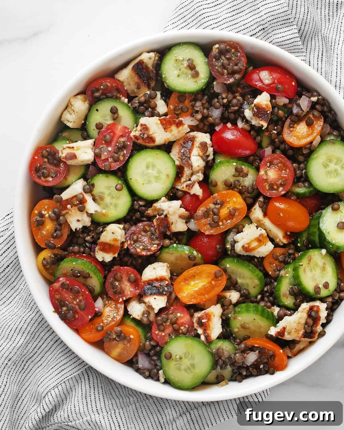 Mediterranean Grilled Halloumi Lentil Salad 2 A large bowl filled with a vibrant grilled halloumi salad, featuring golden-brown halloumi cubes, dark black lentils, sliced green cucumbers, red cherry tomatoes, and finely chopped red onions.