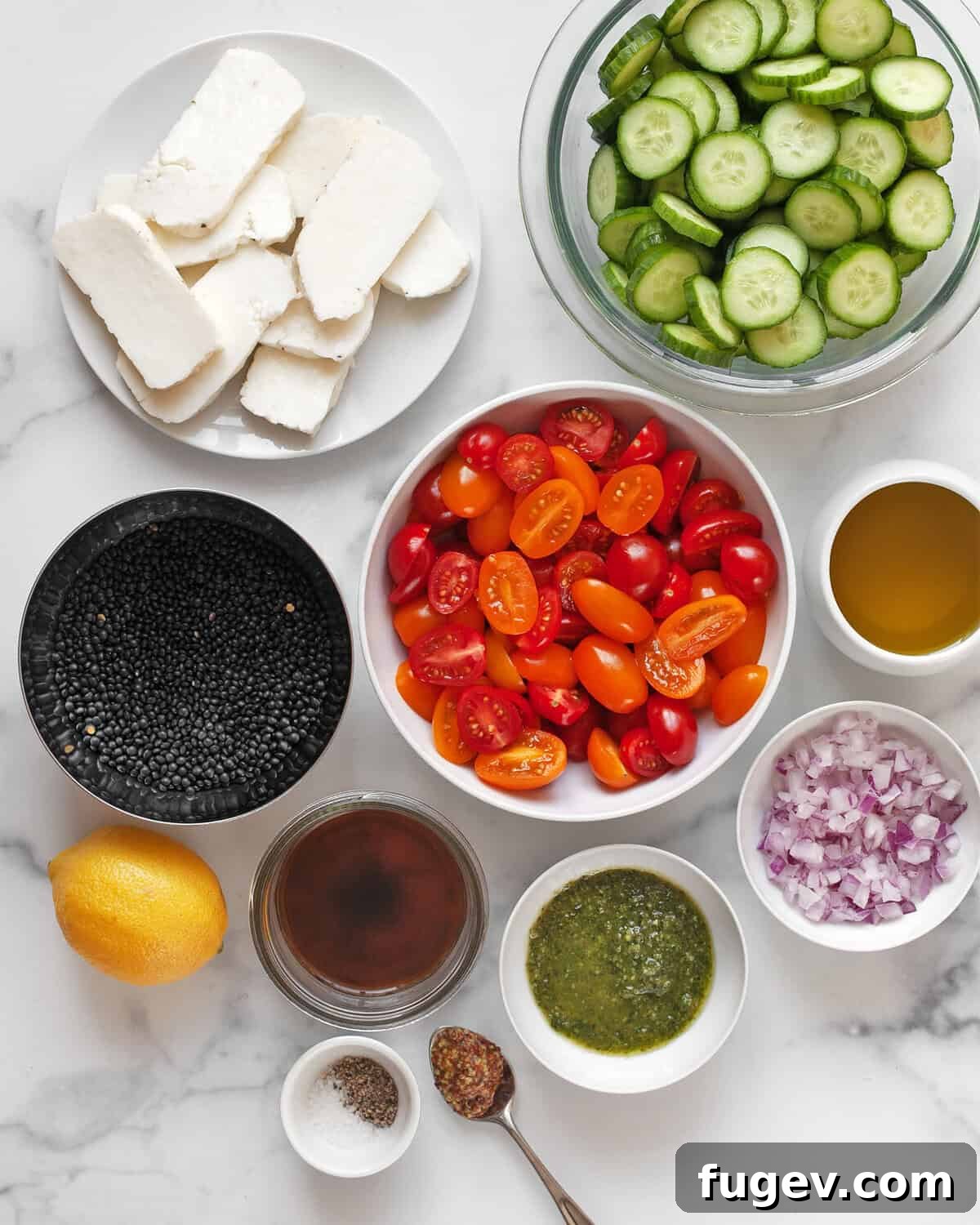 Mediterranean Grilled Halloumi Lentil Salad 3 Arrangement of fresh ingredients for grilled halloumi lentil salad: blocks of halloumi cheese, a bowl of black lentils, halved cherry tomatoes, sliced cucumbers, red onions, a jar of pesto, olive oil, lemon, red wine vinegar, and mustard.