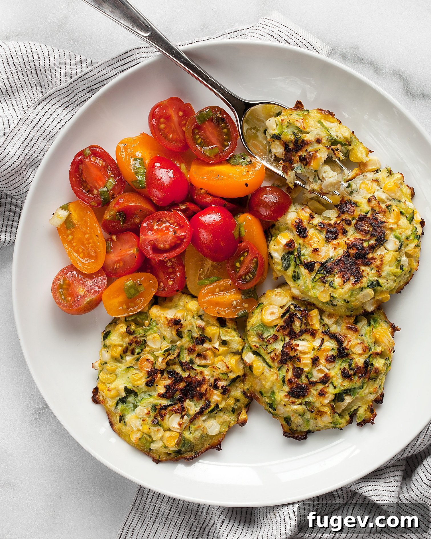 Golden Baked Zucchini Corn Fritters 7 Baked zucchini corn fritters with a tomato salad on a plate, featuring a fork ready to enjoy.