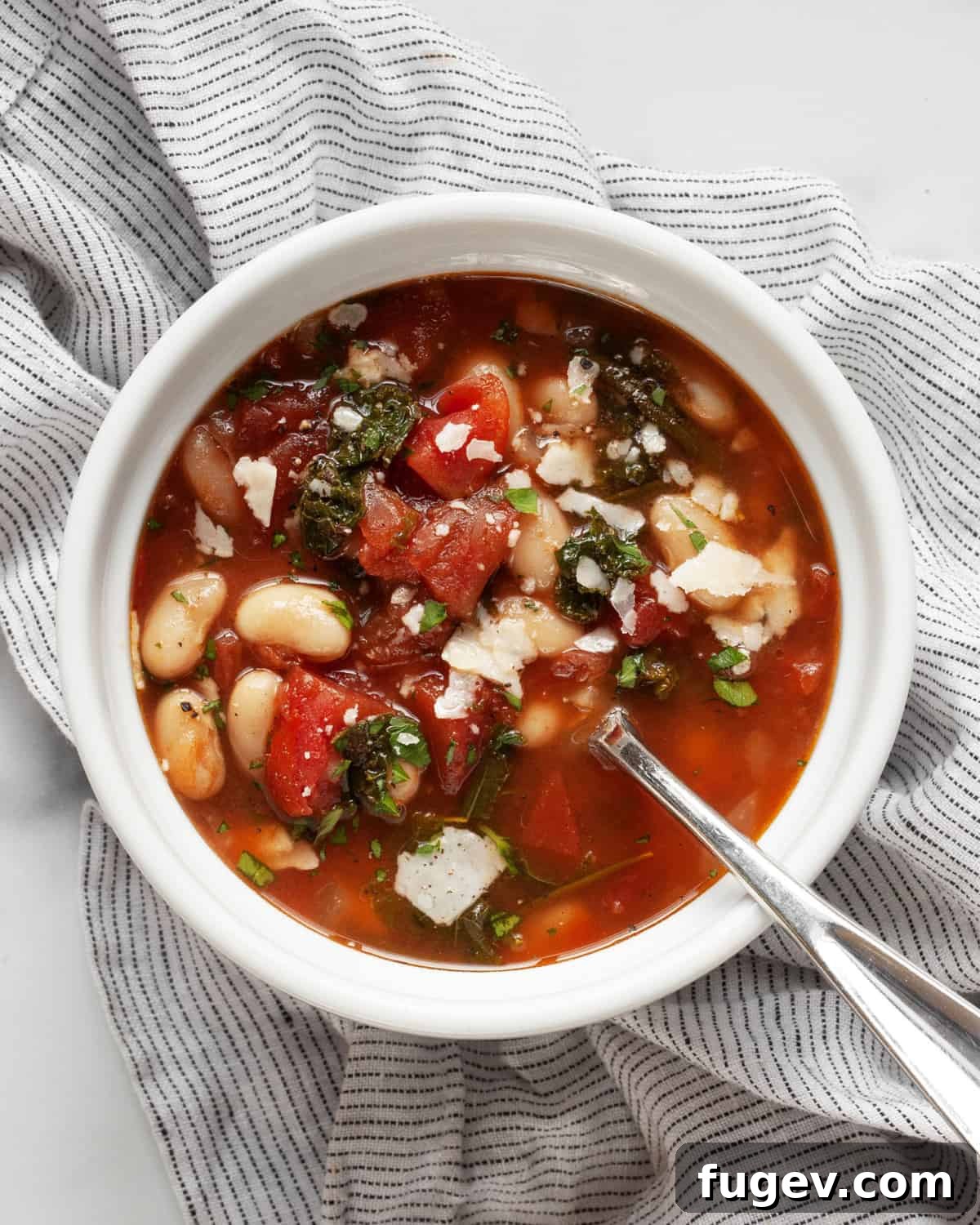 Rustic Rosemary White Bean Tomato Soup 6 A close-up of a single bowl of white bean soup with tomatoes and kale, garnished with fresh herbs and a spoon.