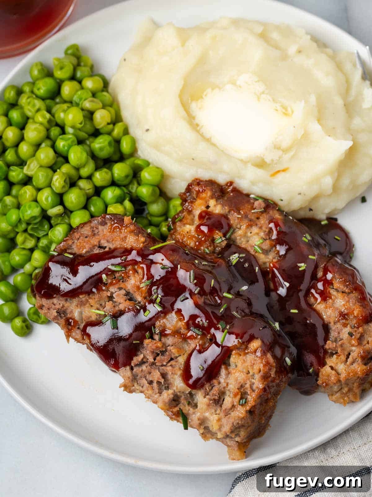 Sticky-Sweet Brown Sugar Meatloaf 2 Sliced brown sugar glazed meatloaf on a plate with creamy mashed potatoes and green peas, showcasing its moist texture and rich glaze.