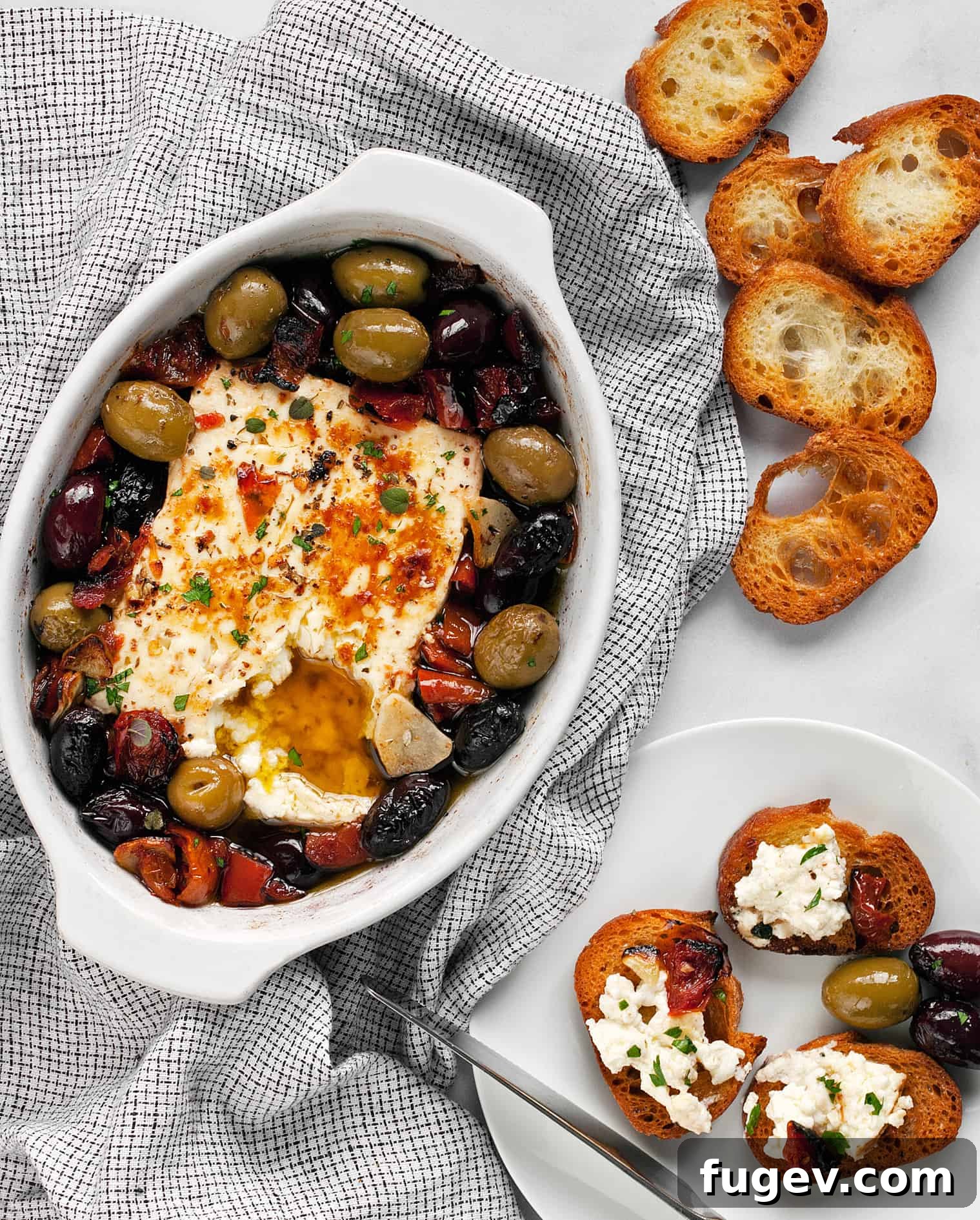 Roasted Feta with Burst Tomatoes and Olives 2 Baked feta with tomatoes and olive in a dish with toasted baguette slices.