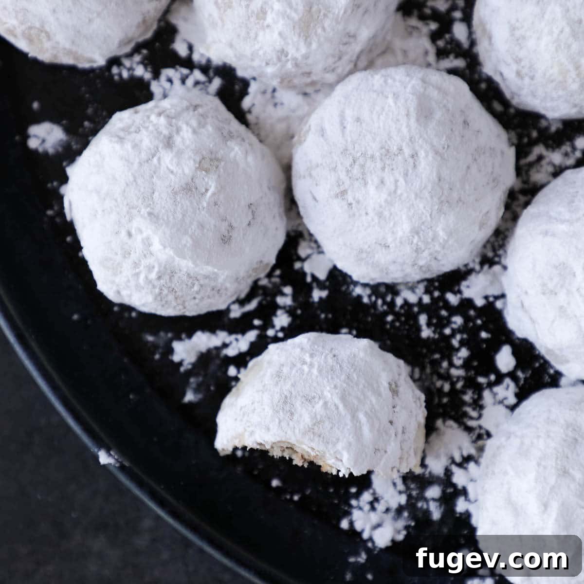 Melt-in-Your-Mouth Snowball Cookies 3 A group of white snowball cookies on a black plate, perfectly dusted with powdered sugar.