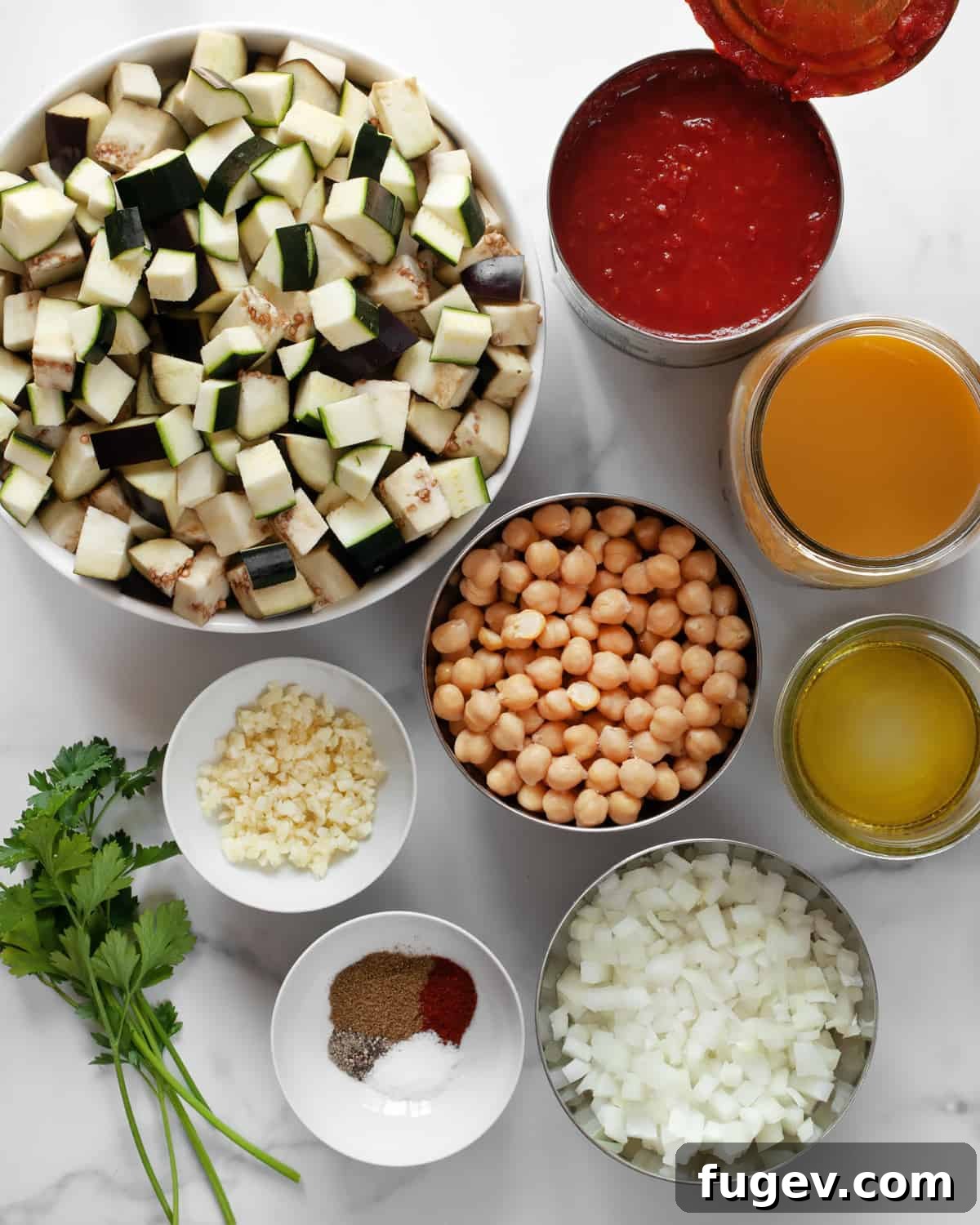 Mediterranean Eggplant and Chickpea Stew 3 A vibrant selection of fresh ingredients including whole eggplants, zucchini, a can of crushed tomatoes, chickpeas, onions, garlic cloves, various spices, olive oil, and vegetable broth, neatly laid out on a kitchen counter.