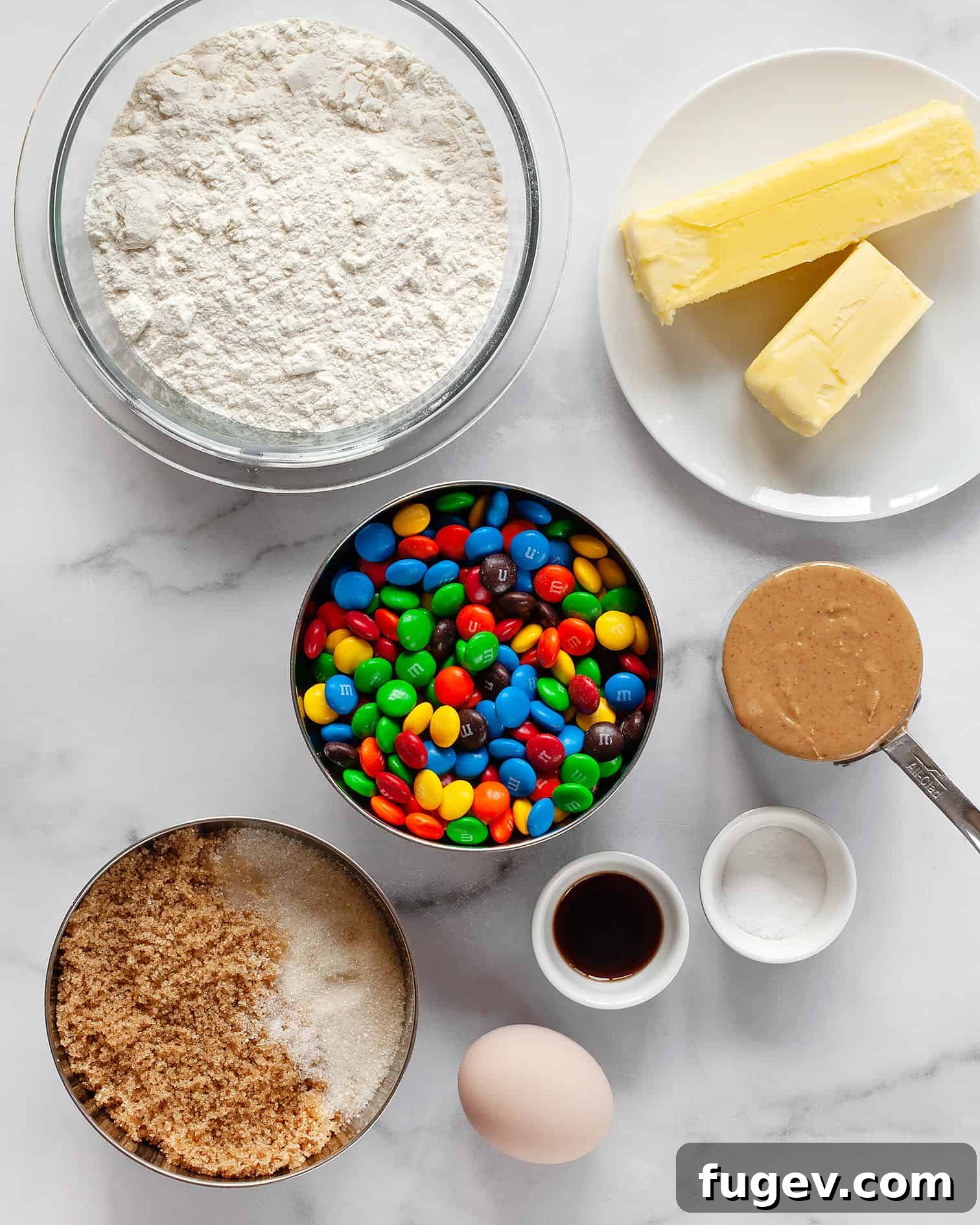 Chewy Peanut Butter Chocolate Candy Cookies 3 A flat lay image displaying all the key ingredients for Peanut Butter M&M cookies: M&Ms, flour, creamy peanut butter, unsalted butter, vanilla extract, and an egg.