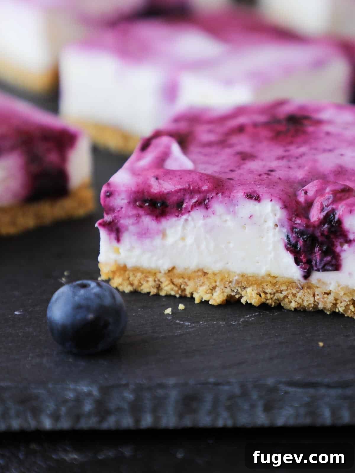 Refreshing No Bake Blueberry Cheesecake Squares 5 A fresh blueberry next to a square no-bake blueberry cheesecake bar, highlighting the vibrant fruit and creamy dessert.