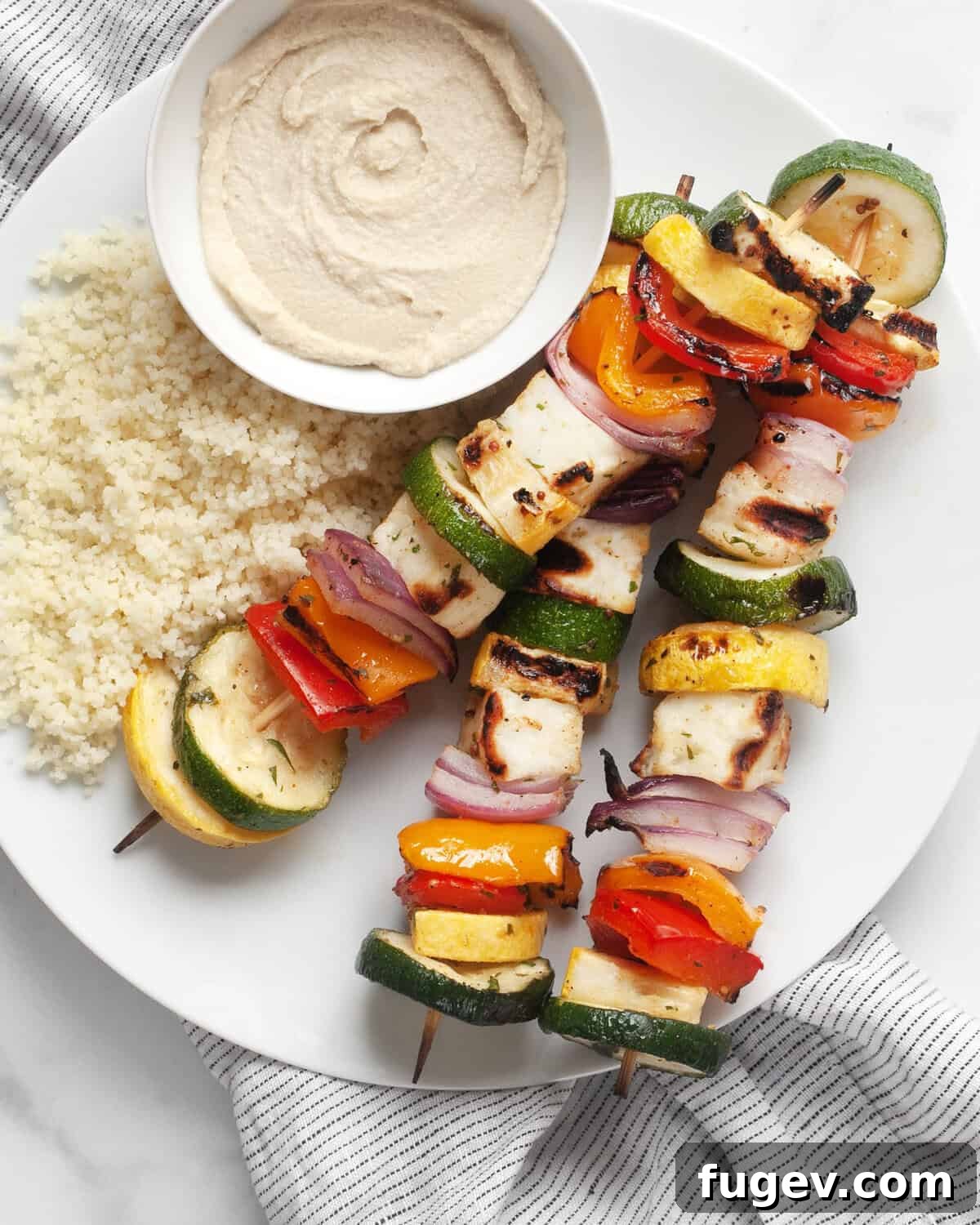 Golden Halloumi Harvest Skewers 4 Three grilled veggie and halloumi skewers artfully arranged on a plate with fluffy couscous and creamy hummus.