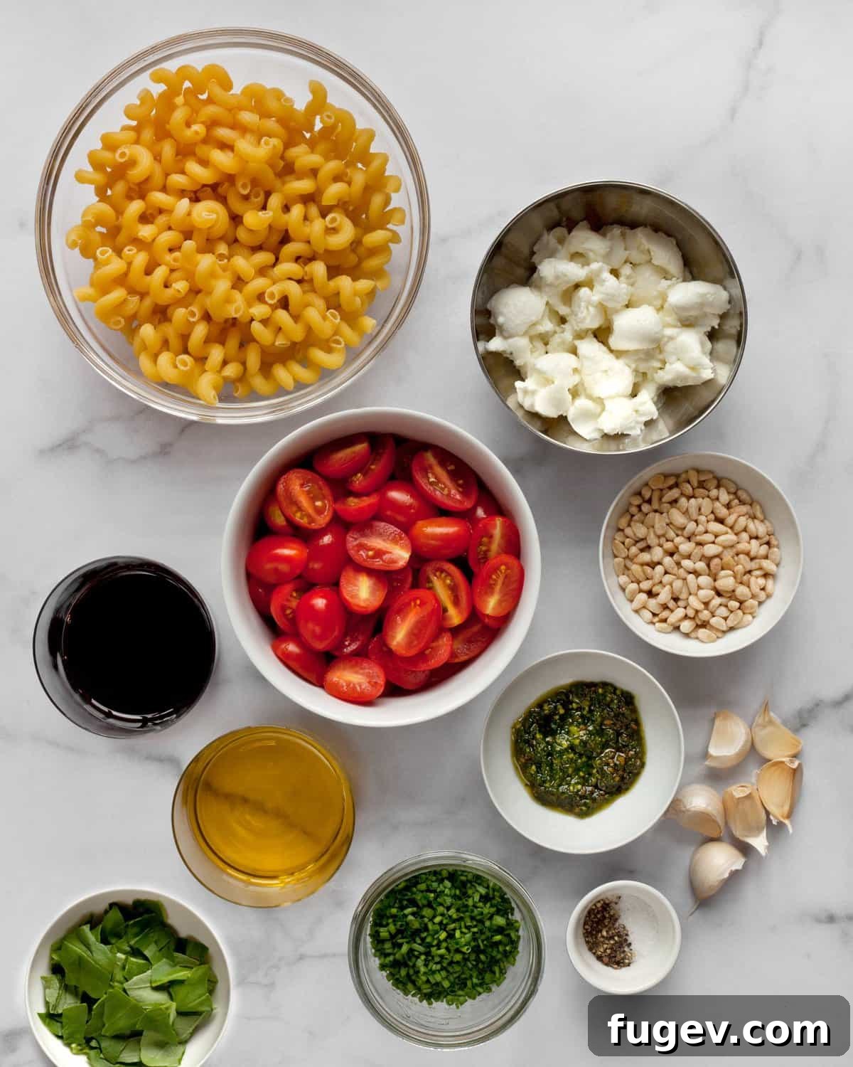 Sun-Kissed Tomato Pesto Pasta Salad 4 Ingredients for pesto pasta salad including pasta, halved tomatoes, fresh mozzarella, basil, a jar of pesto, olive oil, and whole garlic cloves.