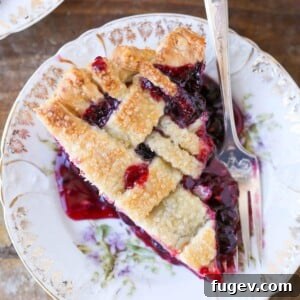A perfectly cut serving of Mixed Berry Pie, showcasing its vibrant berry filling beneath a beautifully woven lattice top, ready to be enjoyed with a silver fork.