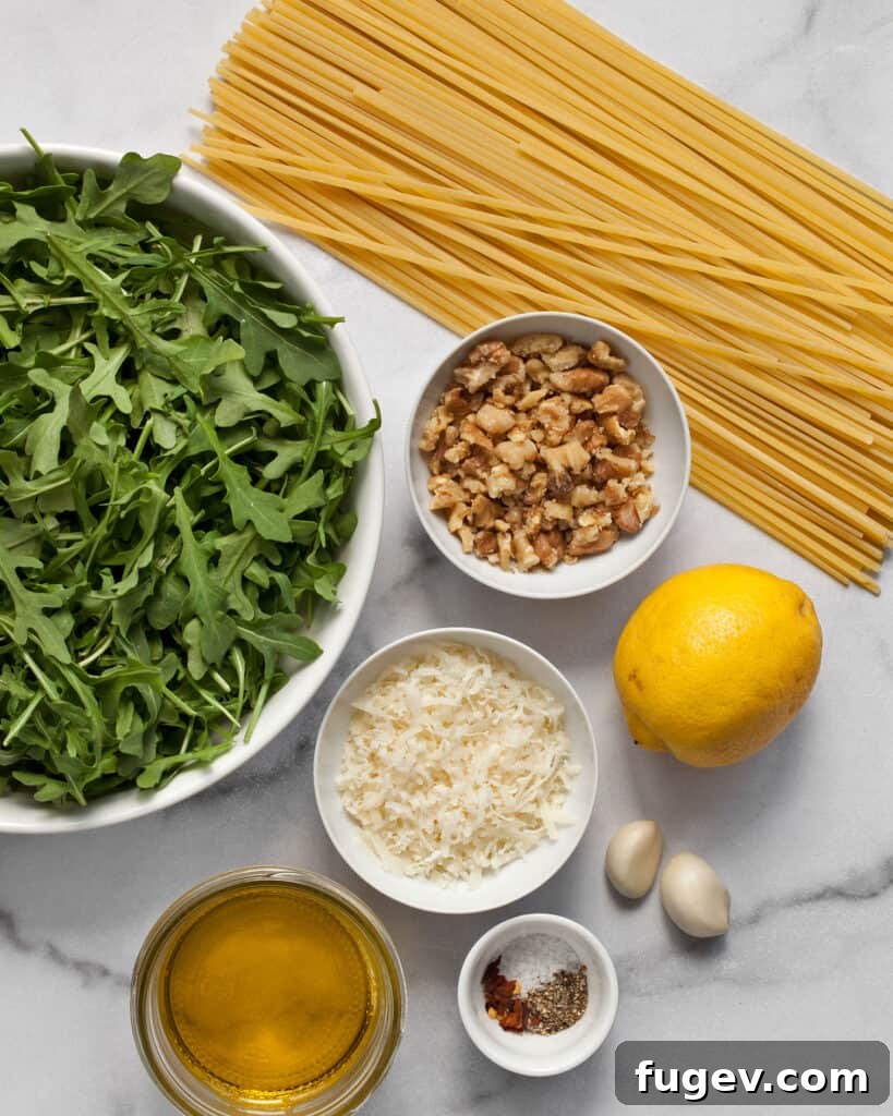 Peppery Arugula Pesto Pasta 3 A selection of fresh ingredients including dry pasta, a bunch of arugula, a bright lemon, garlic cloves, a bottle of olive oil, and a small bowl of walnuts, all ready for the recipe.