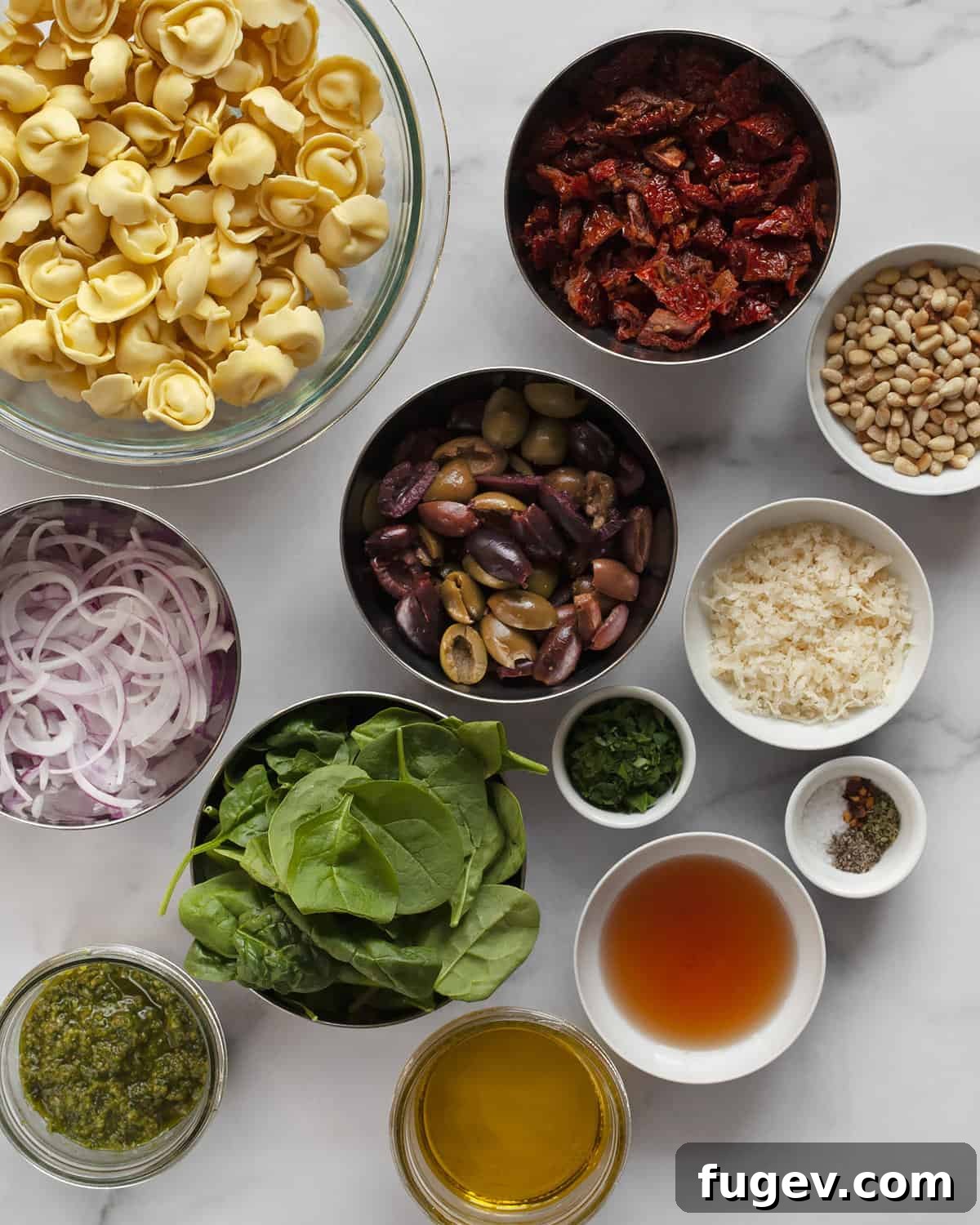 Vibrant Tortellini Salad 3 An array of fresh ingredients for the pasta salad including tortellini, sun-dried tomatoes, olives, spinach, Parmesan, red onions, pesto, and pine nuts.