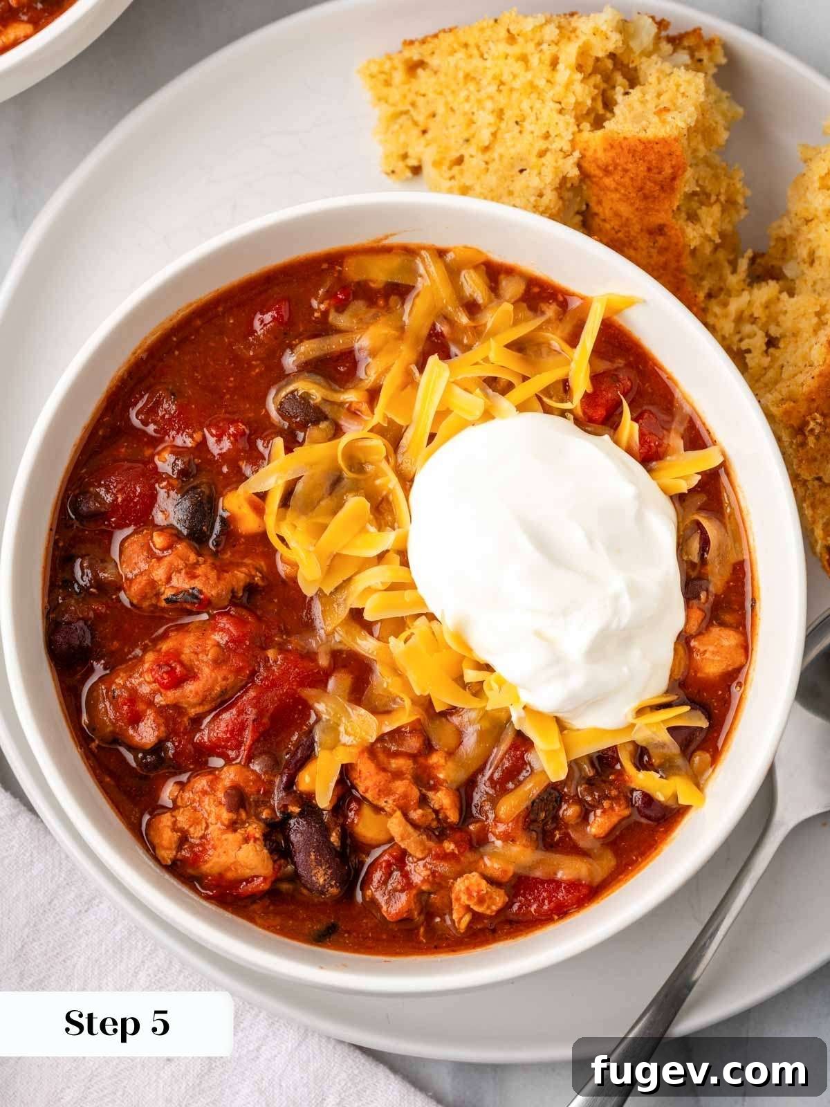 Smoldering Chipotle Chili 9 A close-up of the finished chipotle chili in a white bowl, generously topped with a swirl of sour cream and a mound of shredded cheddar cheese, ready to be enjoyed.