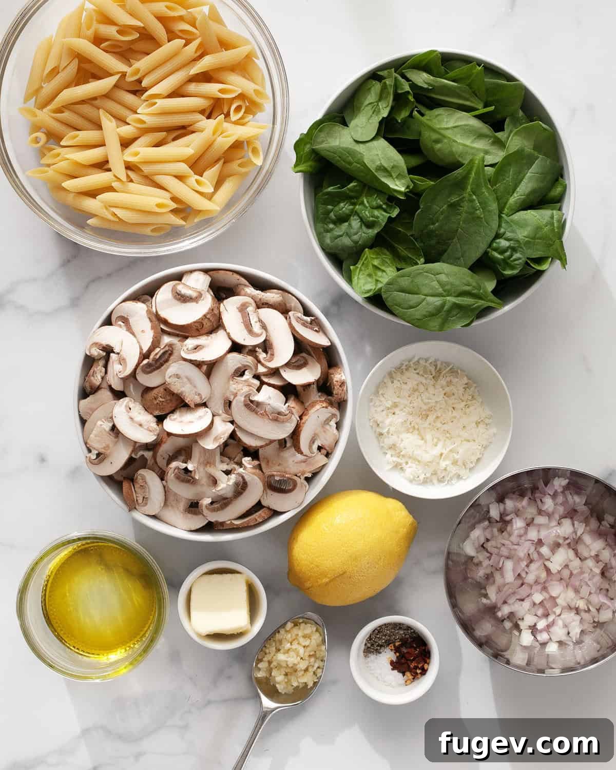 A flat lay photograph showcasing all the fresh ingredients needed for mushroom spinach pasta, including sliced mushrooms, fresh spinach leaves, penne pasta, lemons, garlic, olive oil, Parmesan cheese, shallots, butter, and various seasonings.
