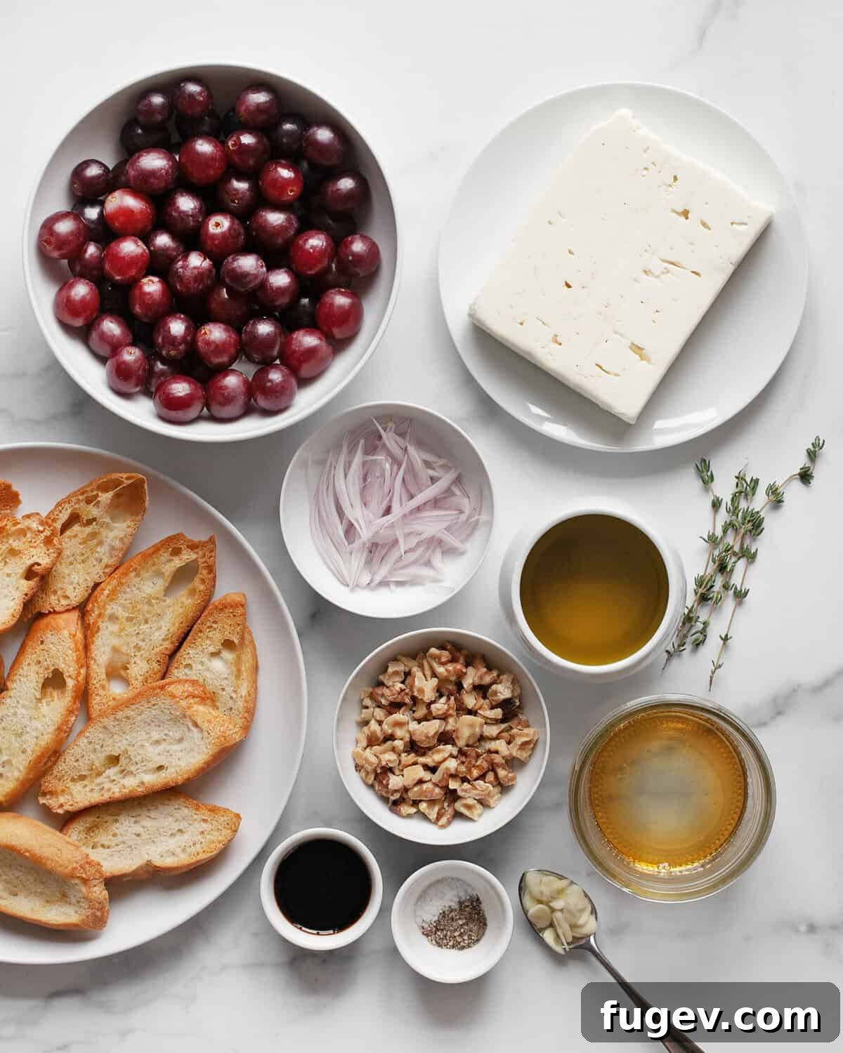Golden Baked Feta with Honey-Kissed Grapes 3 A flat lay of ingredients for baked feta and grapes, including a block of feta, red and green grapes, a bottle of olive oil, balsamic vinegar, sliced shallots, garlic, thyme sprigs, walnuts, salt, pepper, and toasted bread slices.
