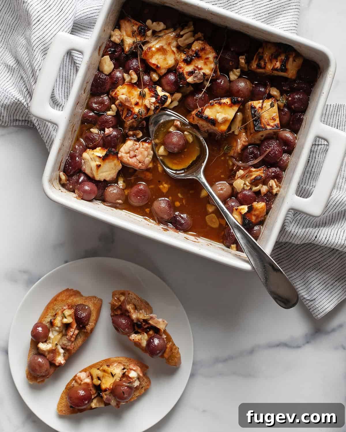 Golden Baked Feta with Honey-Kissed Grapes 6 A baking dish of baked feta and grapes with a serving spoon, beside a plate with three crostini topped with feta, grapes, and walnuts.