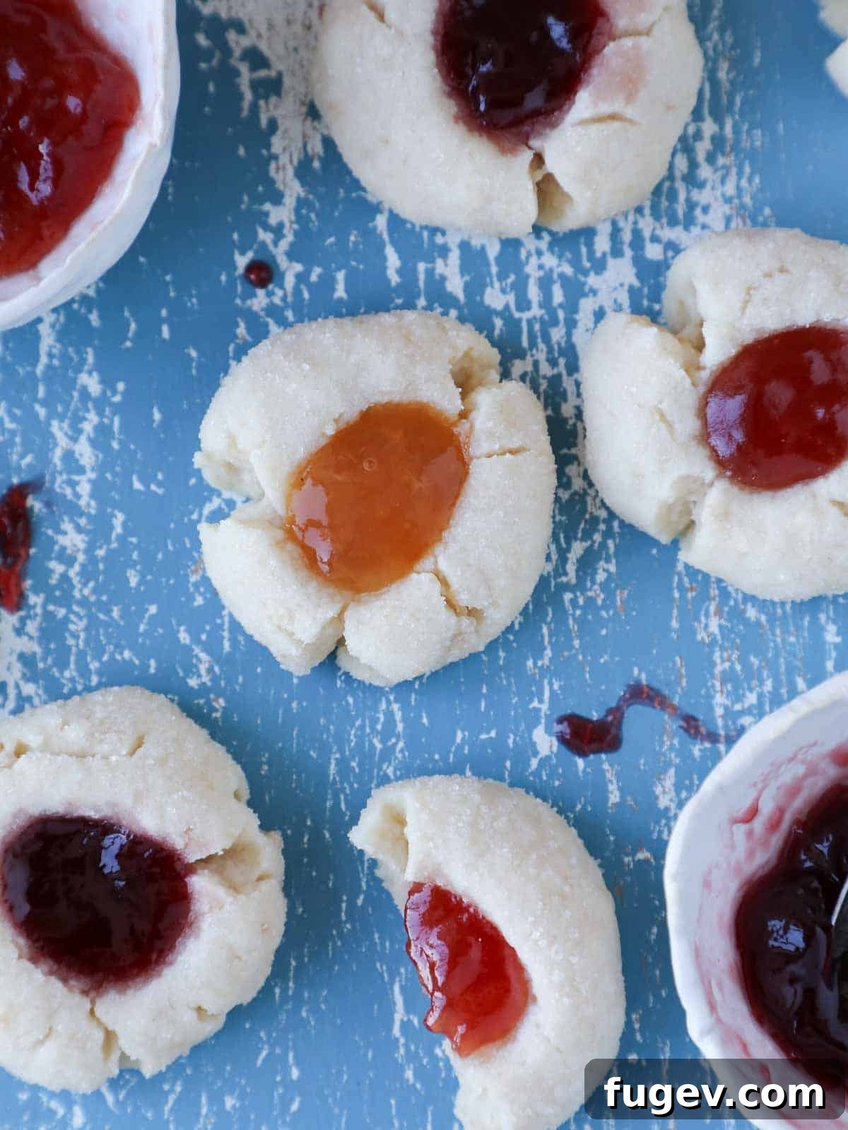 Perfectly Jammed Thumbprints 2 A collection of beautifully baked thumbprint cookies, some filled with strawberry jam, others with apricot, arranged on a rustic blue board alongside jars of various jams, suggesting their delightful filling options. The image highlights the golden edges and colorful centers of the cookies.