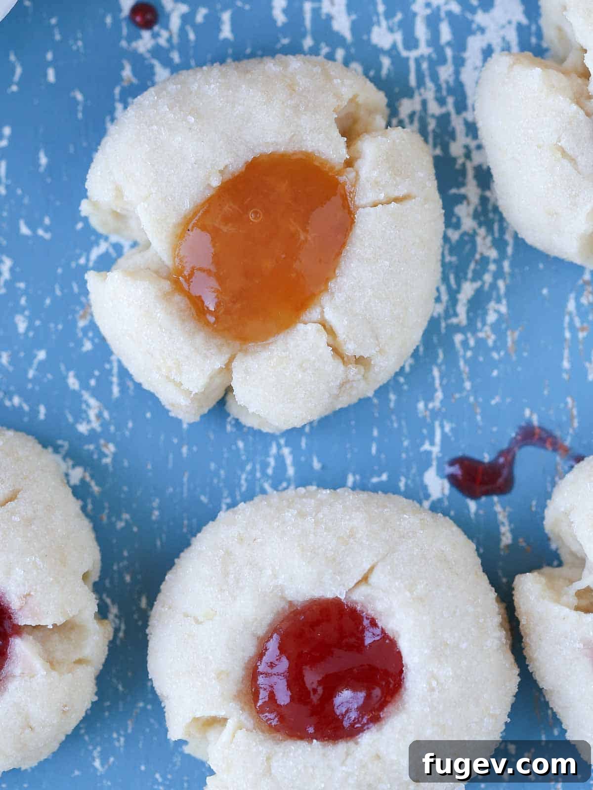 Perfectly Jammed Thumbprints 11 Two perfectly baked thumbprint cookies, one with strawberry jam and one with apricot jam, resting on a smooth blue surface, showcasing their pristine appearance.