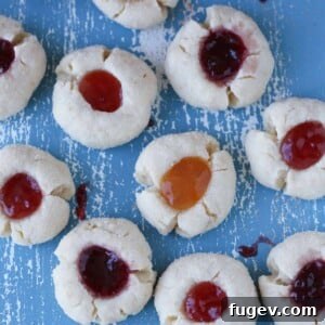 A collection of beautifully baked thumbprint cookies, some filled with strawberry jam, others with apricot, arranged on a rustic blue board alongside jars of various jams, suggesting their delightful filling options.
