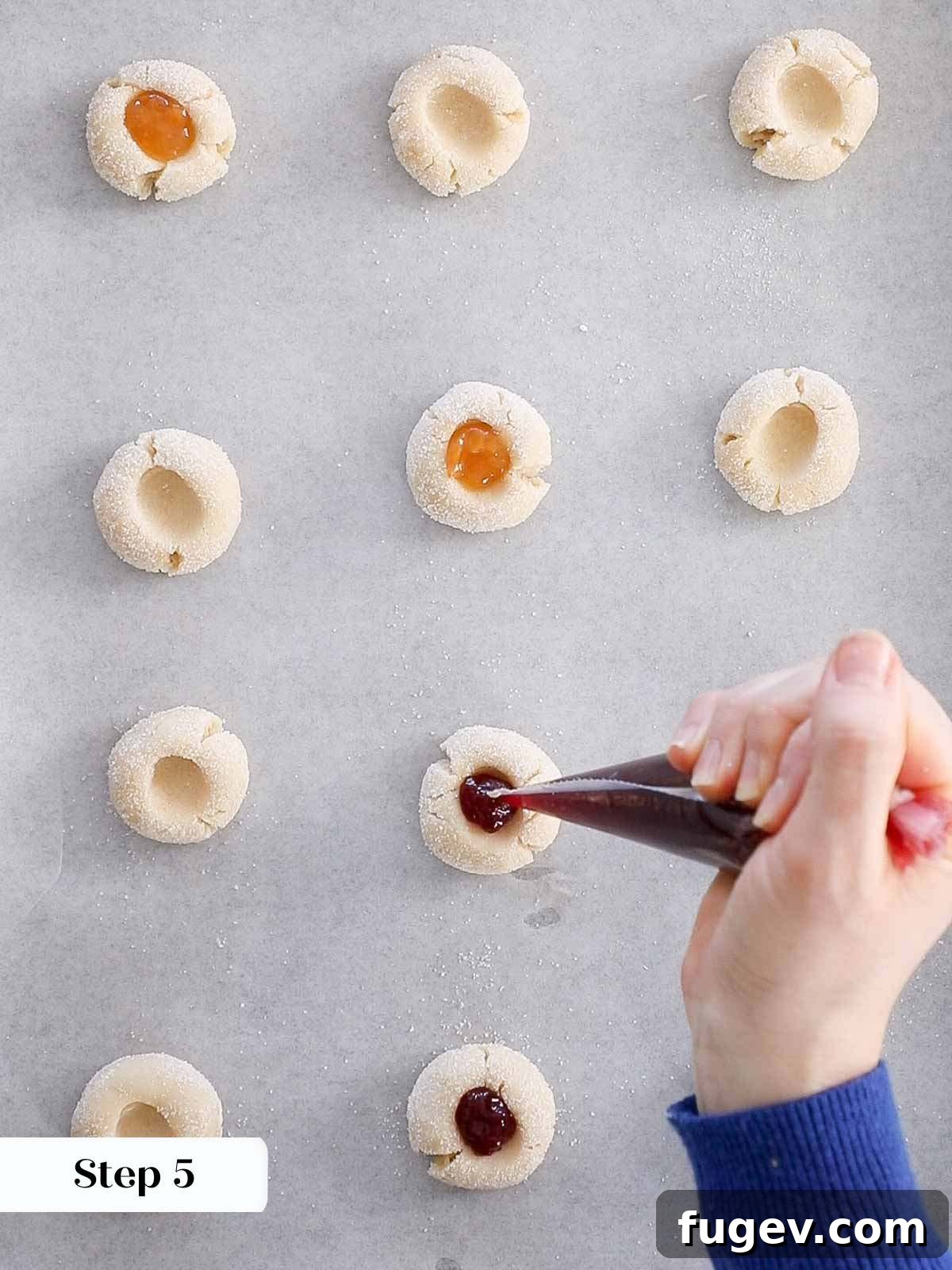 Perfectly Jammed Thumbprints 9 Thumbprint cookie dough balls on a baking sheet, each indentation meticulously filled with colorful jam before going into the oven. The image showcases the variety of jam fillings.