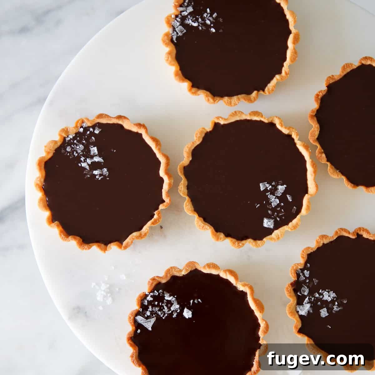 Mastering Chocolate Ganache 5 Elegant mini salted chocolate tarts arranged on a white marble cake stand.