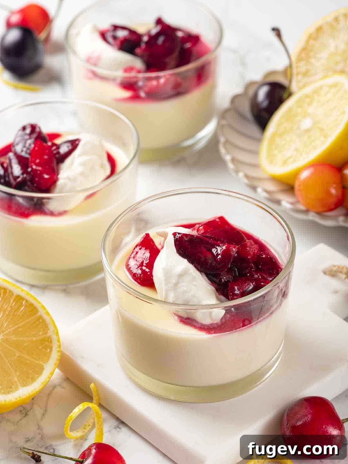 Zesty Lemon Cream 2 Three lemon possets in clear glasses, garnished with whipped cream and a vibrant cherry sauce. The dessert is a smooth, pale yellow, indicating its rich lemon flavor and creamy texture.