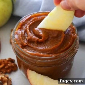 Cozy Spiced Pumpkin Butter 8 A hand scooping thick, spiced pumpkin butter from a glass jar with an apple slice ready for dipping.