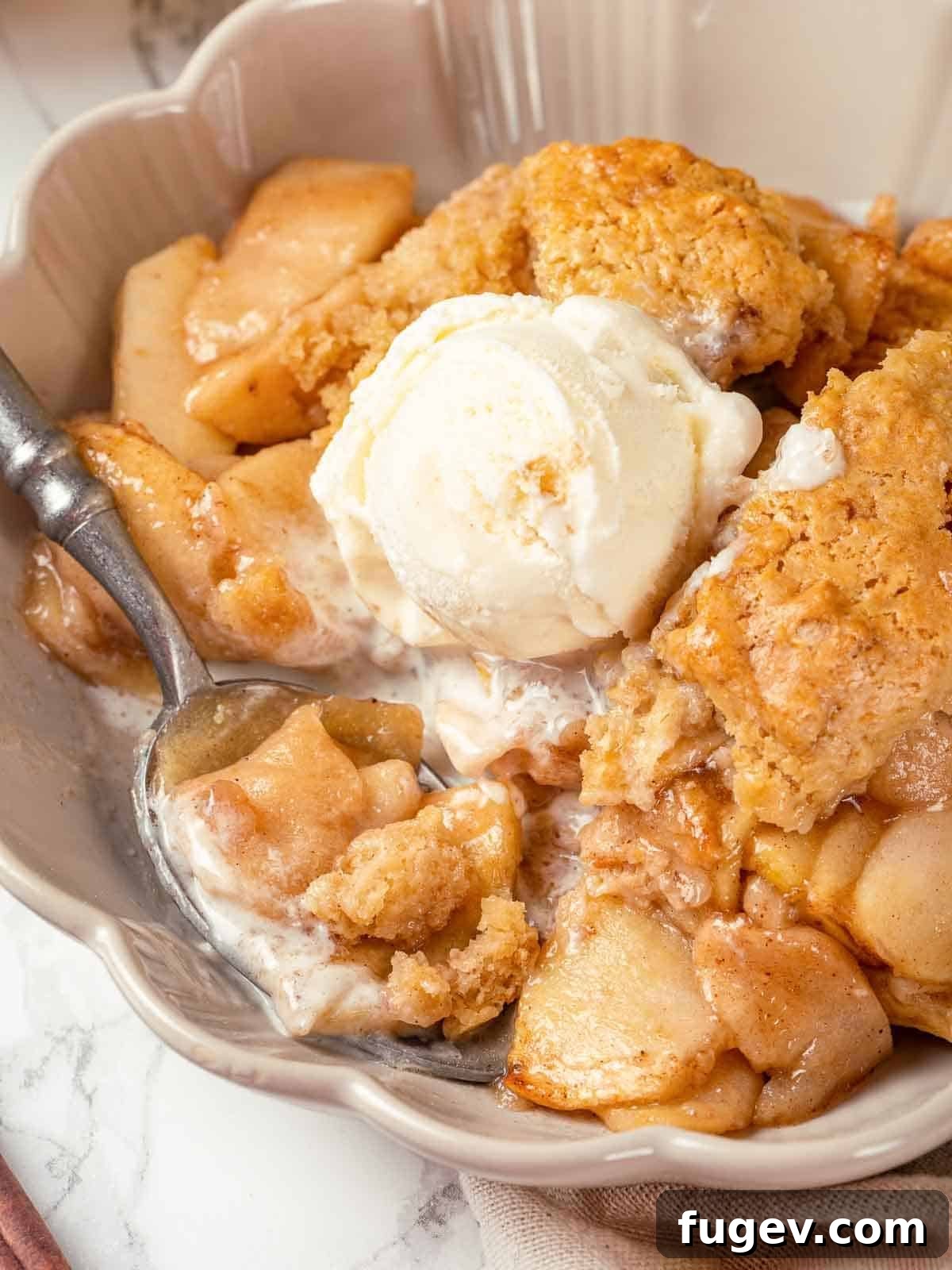 Cozy Apple Cobbler Magic 2 A generous serving of apple cobbler in a white bowl, ready to be enjoyed with a spoon. The golden biscuit topping and sweet apple filling are visible.