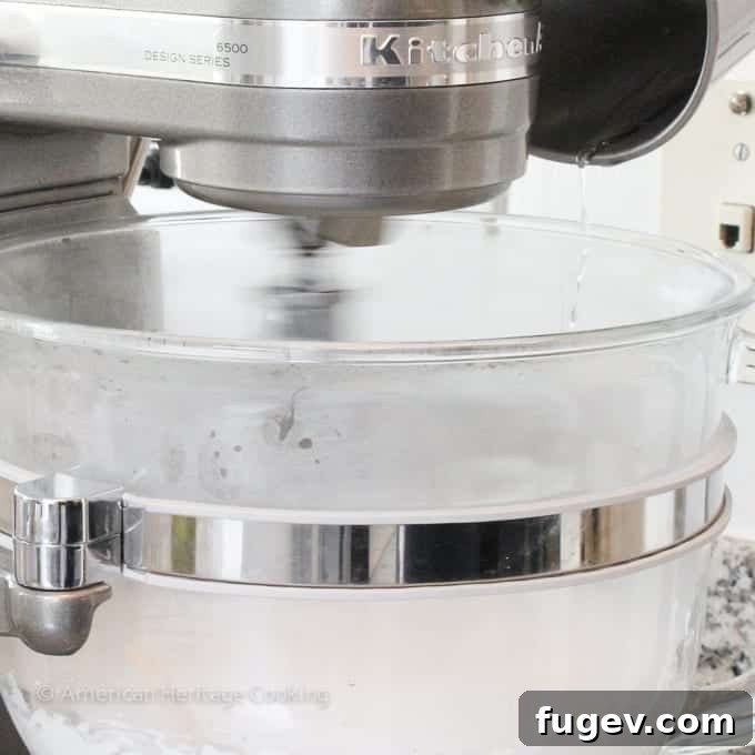 The whisk attachment of the stand mixer blurred in motion, illustrating high-speed whipping as hot sugar syrup is carefully poured down the side of the bowl.