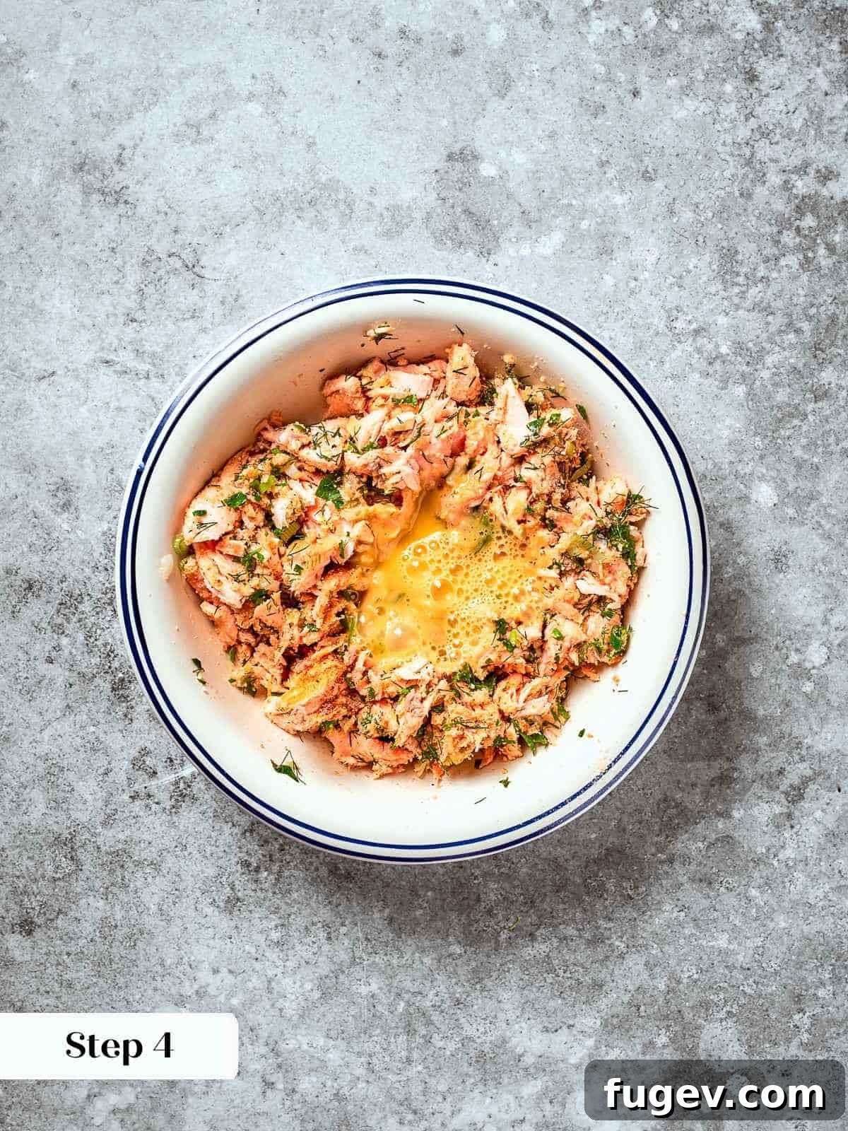 Gourmet Salmon Burgers 7 Egg being added to salmon patty mixture in a mixing bowl with herbs and breadcrumbs.