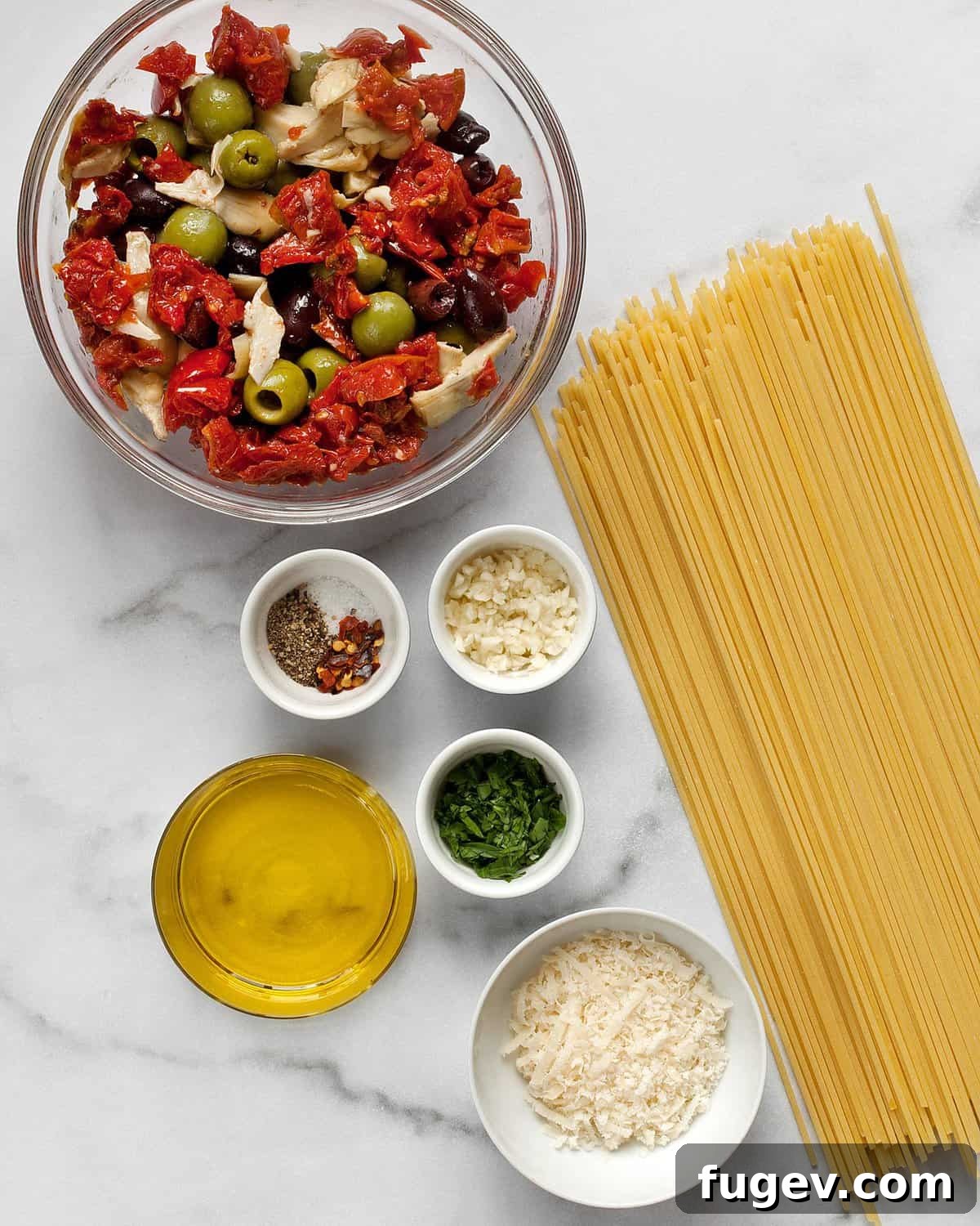 Antipasto Platter Pasta 4 A flat lay of ingredients for antipasto pasta: roasted tomatoes, various olives, artichoke hearts, dry spaghetti, Parmesan cheese, fresh garlic, and olive oil.
