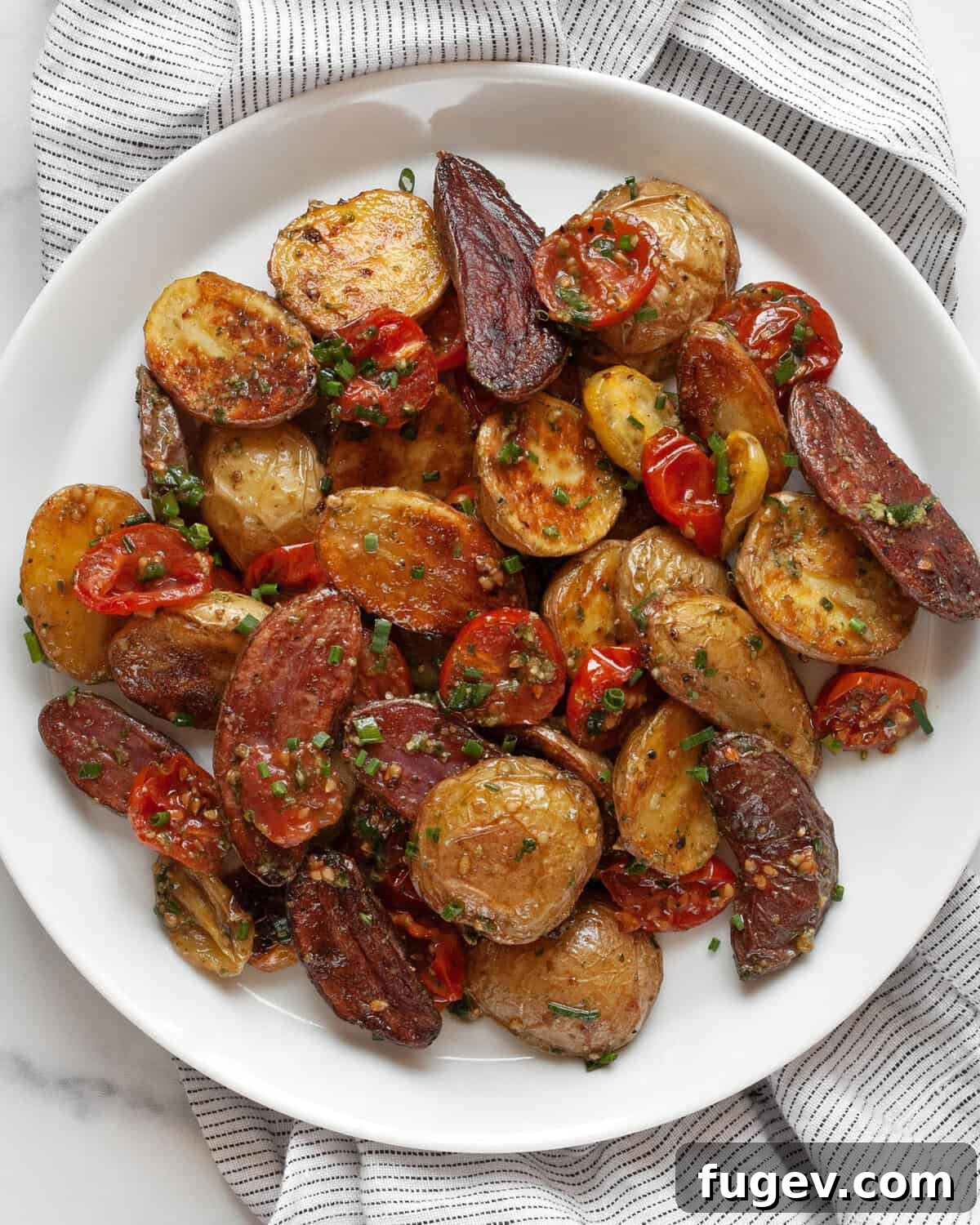 Mediterranean Roasted Tomato Pesto Potato Salad 2 A vibrant serving of roasted tomato pesto potato salad on a white plate, showcasing golden roasted potatoes, bright red blistered tomatoes, and fresh green pesto vinaigrette, garnished with fresh herbs.