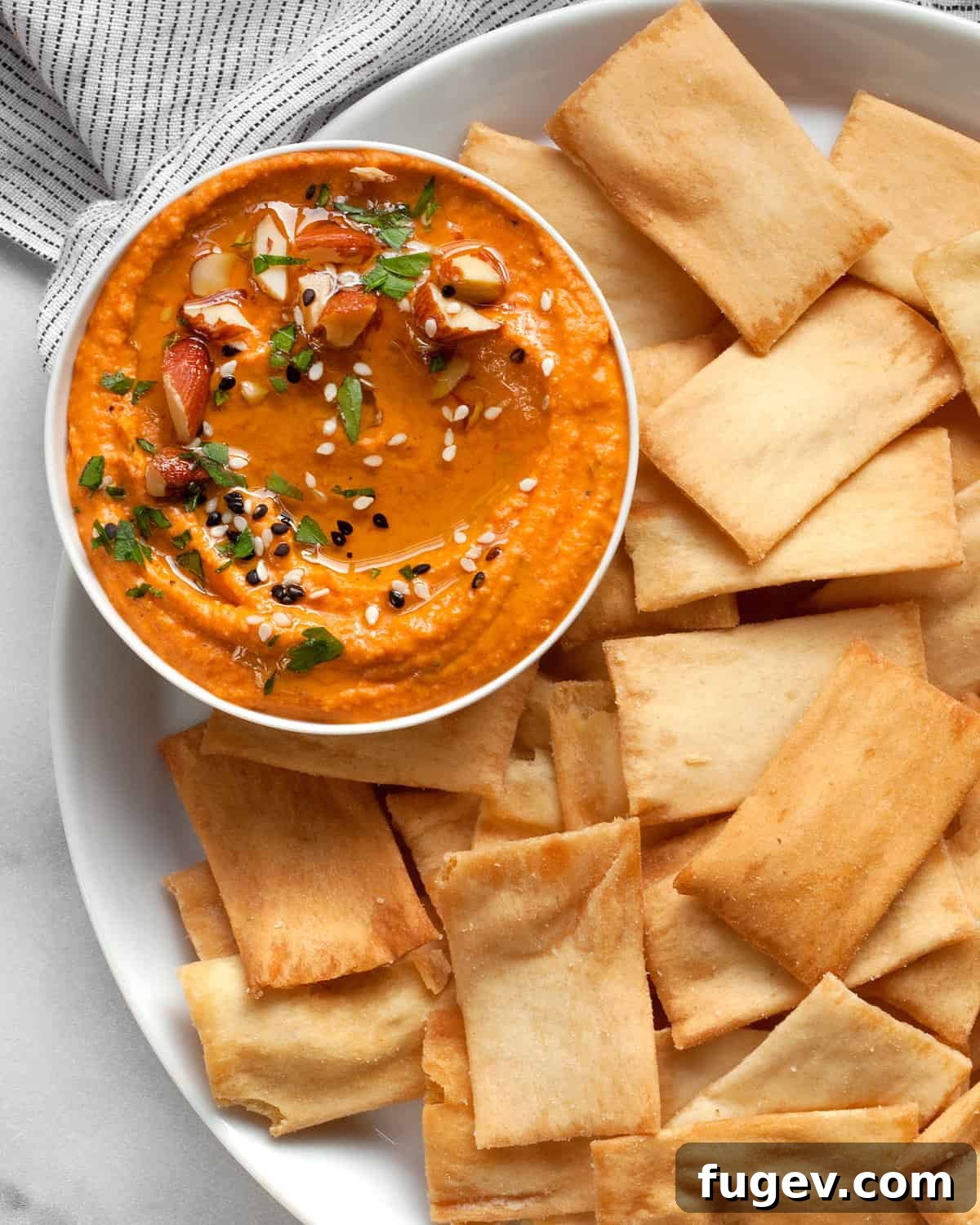 Flame-Kissed Red Pepper Hummus with Smoky Almond Crunch 2 Roasted red pepper hummus in a bowl with pita chips, garnished with olive oil and chopped almonds.