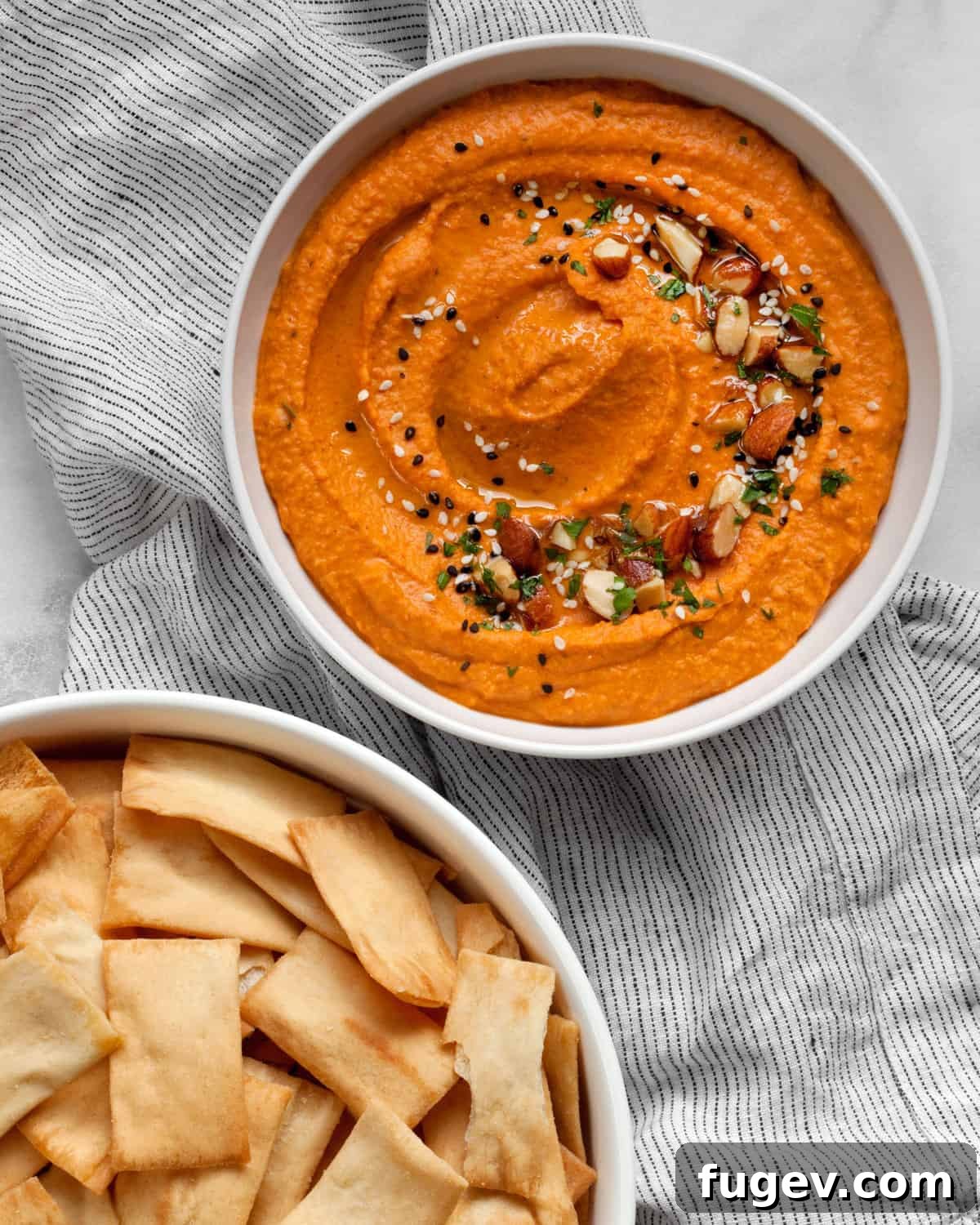 Flame-Kissed Red Pepper Hummus with Smoky Almond Crunch 6 A bowl of red pepper hummus artfully garnished with a swirl of olive oil, chopped almonds, sesame seeds, and fresh parsley, served alongside a separate bowl of crispy pita chips.