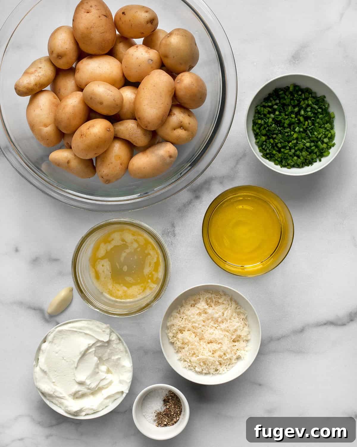 Crispy Accordion Bites 3 A flat lay photograph of all the ingredients laid out: baby potatoes, fresh chives, a bottle of olive oil, a small bowl of melted butter, grated Parmesan cheese, salt, pepper, and a bowl of Greek yogurt.