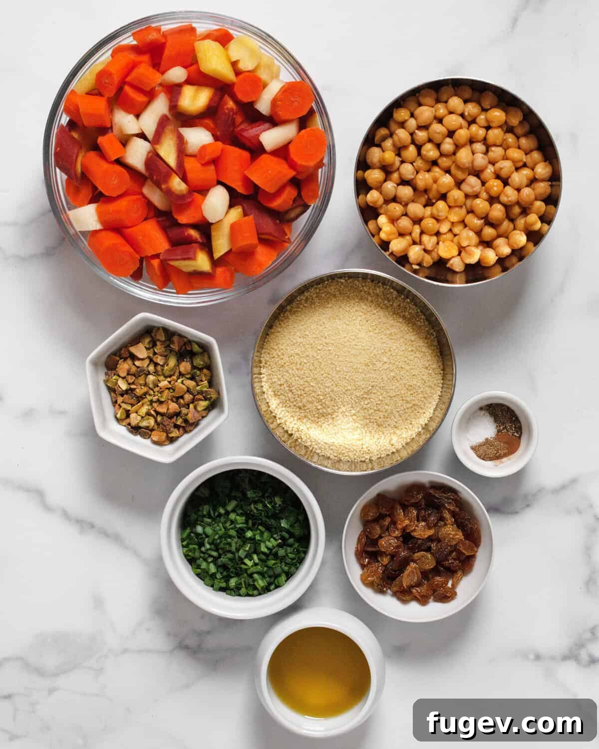 A flat lay photograph showcasing the vibrant ingredients for roasted carrot couscous: carrots, chickpeas, uncooked couscous, golden raisins, fresh parsley, chives, pistachios, various dried spices, and olive oil.