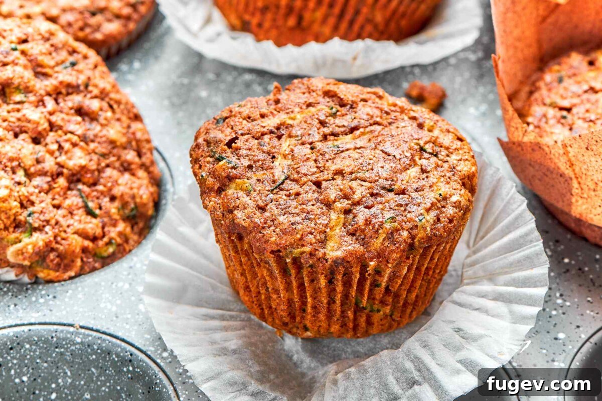 A freshly baked zucchini muffin with its paper wrapper gently peeled down, revealing its moist crumb, tender texture, and visible flecks of green zucchini.
