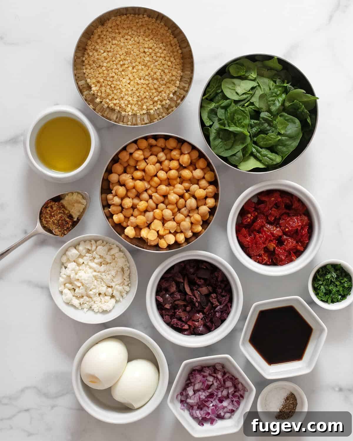 A flat lay photograph showcasing all the fresh and pantry ingredients needed for Mediterranean couscous salad, including pearl couscous, baby spinach, chickpeas, olives, sun dried tomatoes, hard-boiled eggs, feta, olive oil, garlic, mustard, balsamic vinegar, salt and pepper.