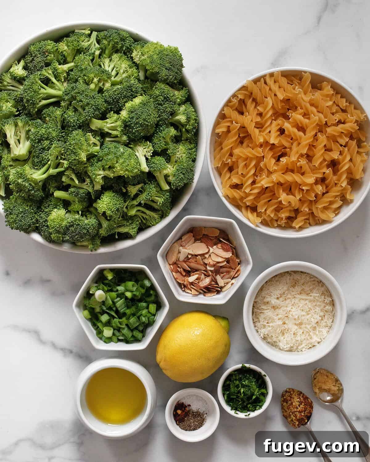 A flat lay photograph showcasing all the fresh ingredients needed for the roasted broccoli pasta salad: broccoli florets, fusilli pasta, sliced almonds, green onions (scallions), grated parmesan cheese, fresh lemons, parsley, garlic, whole grain mustard, olive oil, red pepper flakes, salt, and black pepper.