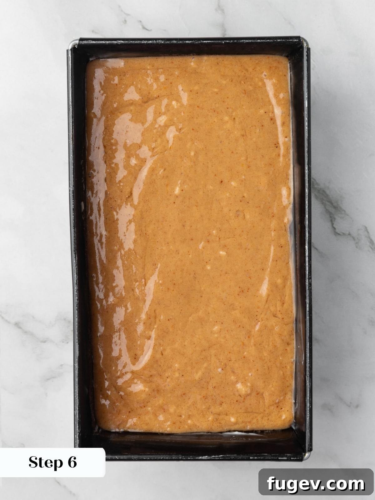 Pale orange batter spread evenly inside a dark metal loaf pan on a marble surface.