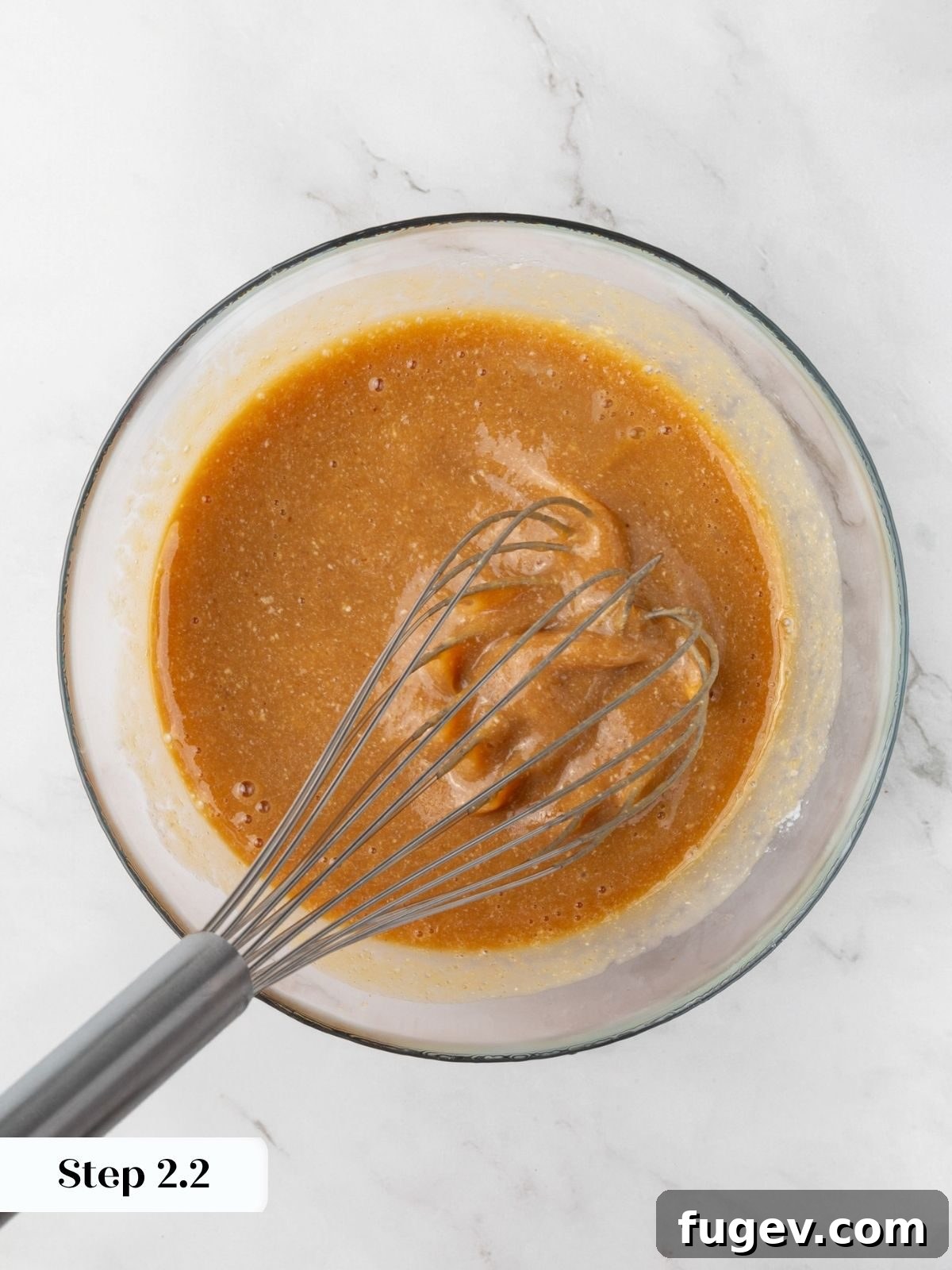 Smooth caramel-colored wet mixture combined evenly in a glass bowl with a metal whisk resting inside.