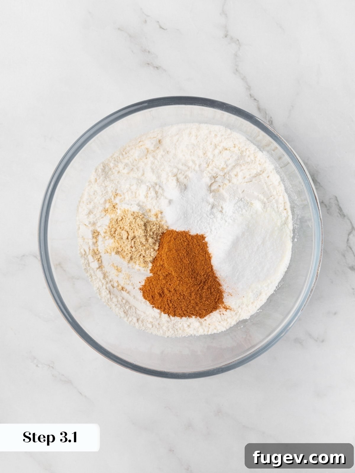 Flour with cinnamon, ginger, and white piles of leavening agents in a clear glass mixing bowl.