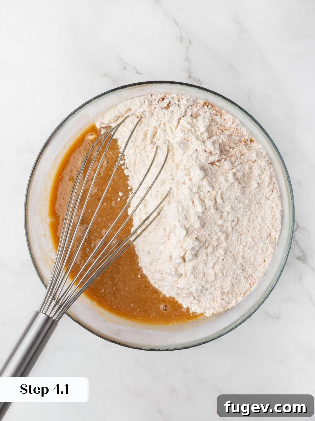 A mound of dry ingredients added atop the caramel-toned wet mixture in a glass mixing bowl.
